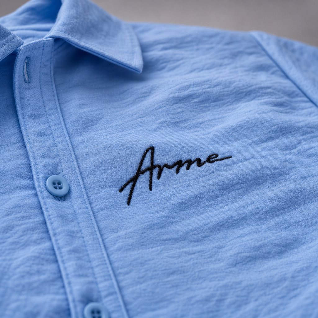 Close-up of black embroidered ARME script logo on the baby blue button-up shirt