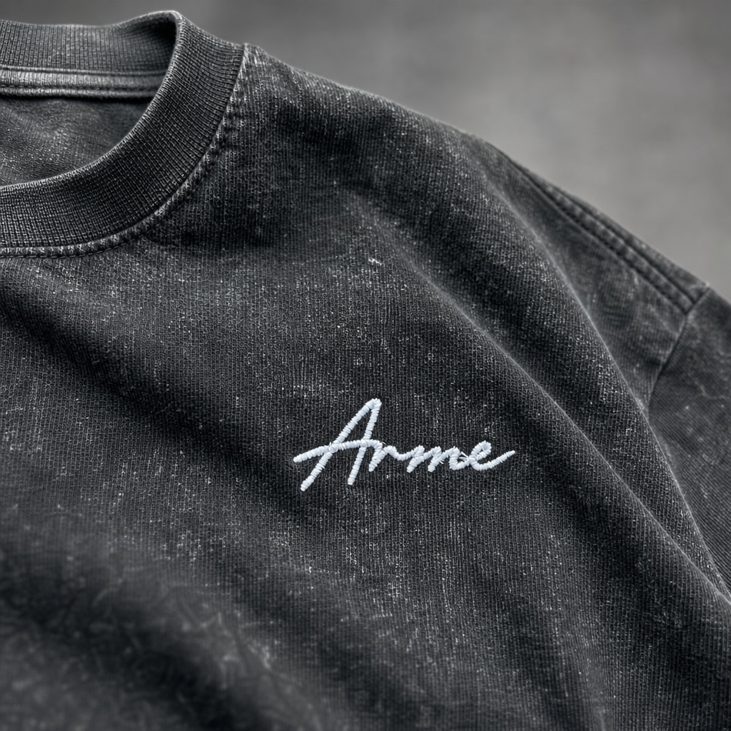 Close-up of white embroidered ARME script logo on washed charcoal cotton tee